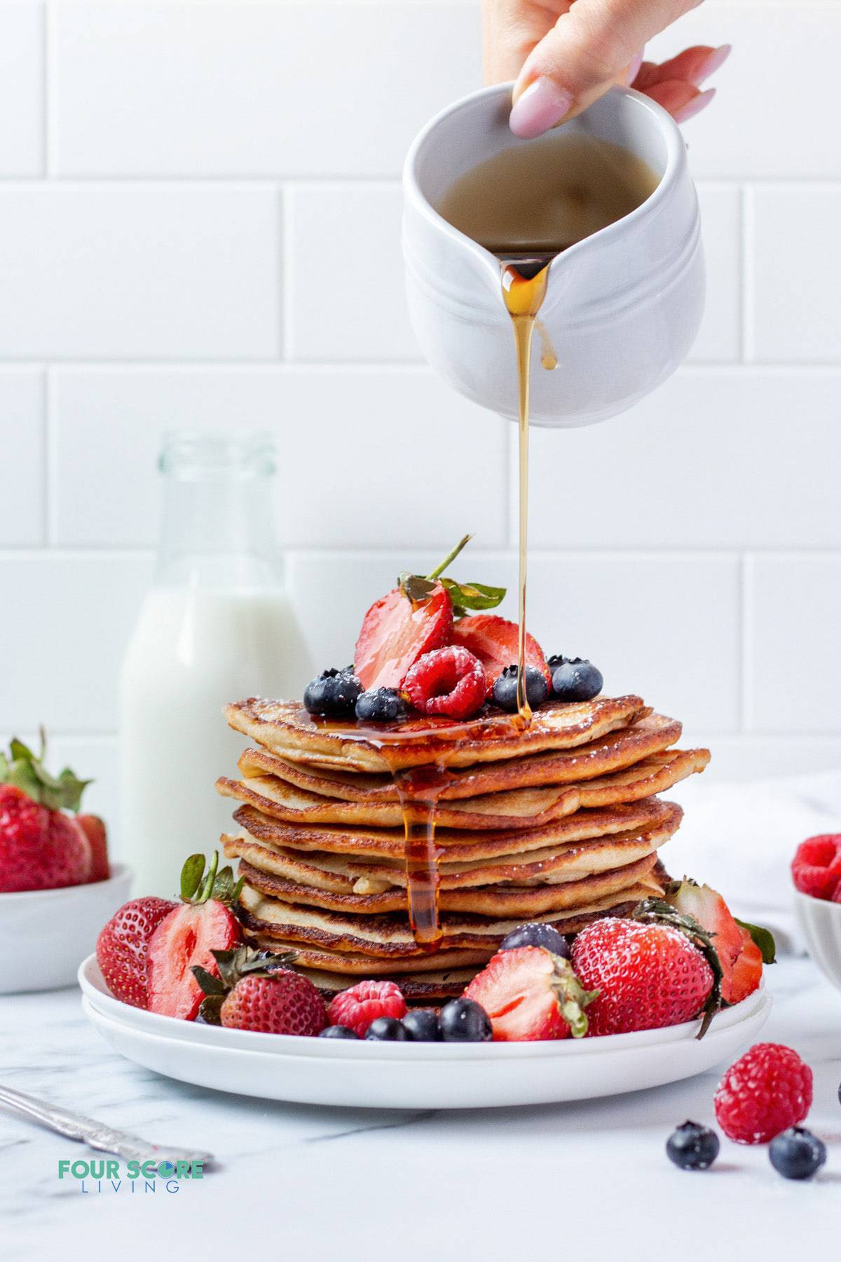 A tall stack of almond flour banana pancakes topped with fresh mixed berries and surrounded by fresh mixed berries. A hand is pouring syrup over the stack.