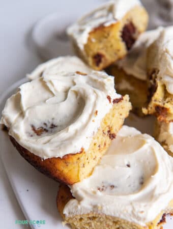 Two cinnamon rolls topped with cream cheese frosting on a plate. More cinnamon rolls in the background.