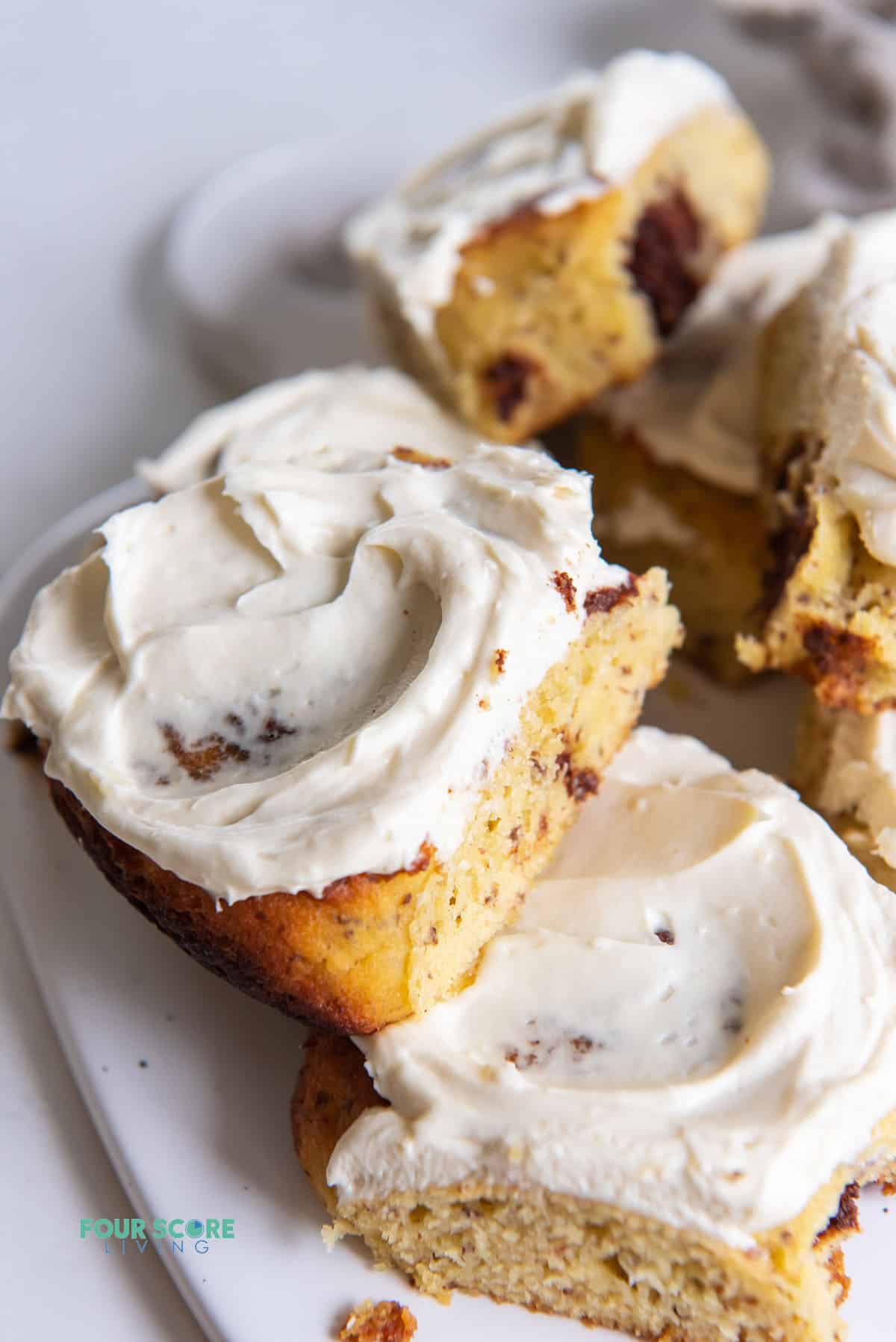 Two cinnamon rolls topped with cream cheese frosting on a plate. More cinnamon rolls in the background.