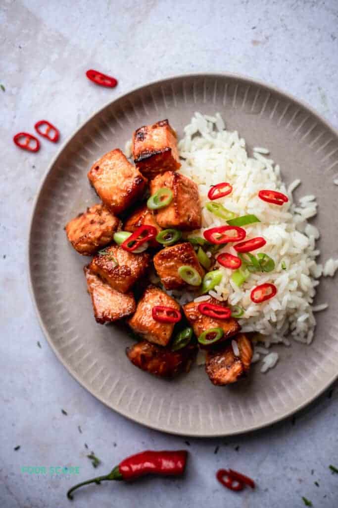 Salmon bites on a plate next to rice with scallions and red peppers scattered on top.