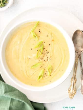 Top view photo of Magic Leek Soup in a white bowl, on a white plate. There are two silver spoons on the plate and a green kitchen towel by the bowl.