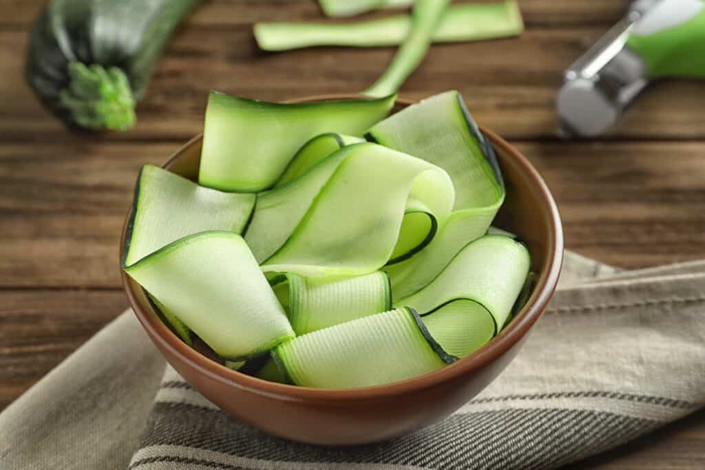 Bowl placed on top of dish towel with wide zucchini noodle slices sitting on a wood table surface.