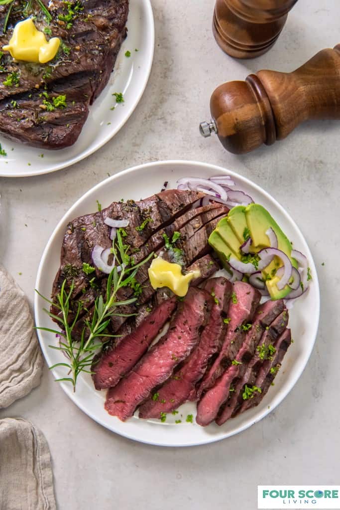 Diagonal, close up image of cooked
and sliced sirloin tip steak on a white dish with a pat of butter, fresh green herbs, a spring of rosemary, sliced ripe avocado and sliced red onion with wooden salt and pepper grinders and a whole, cooked sirloin steak in soft focus in the background.