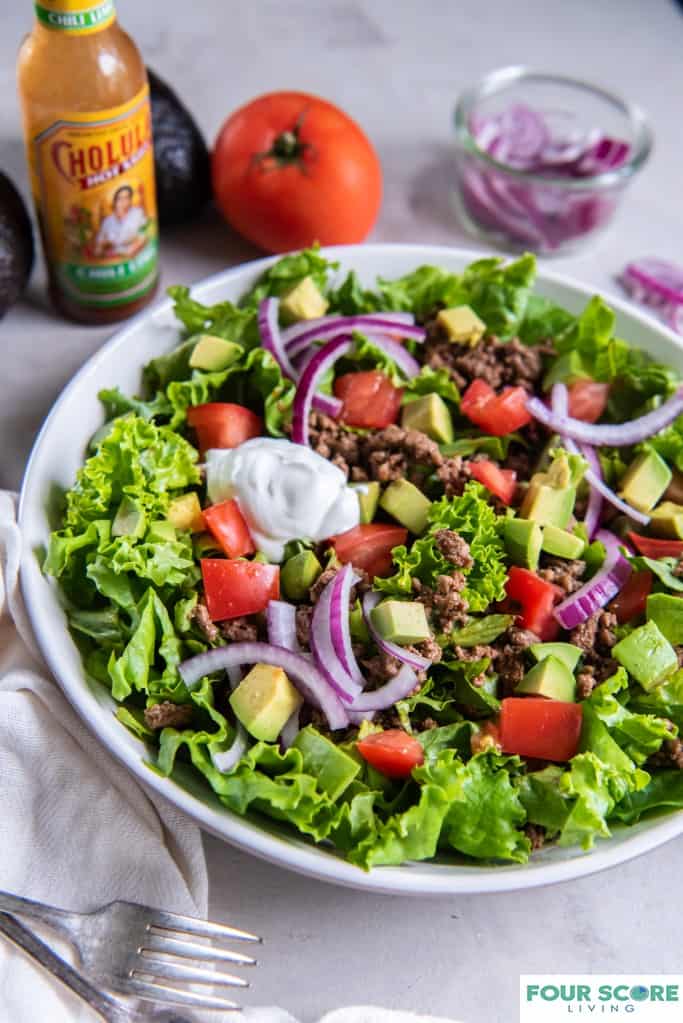 Taco salad in a bowl with chopped tomatoes, sliced red onion, diced avocado, cooked ground beef and a dollop of sour cream.