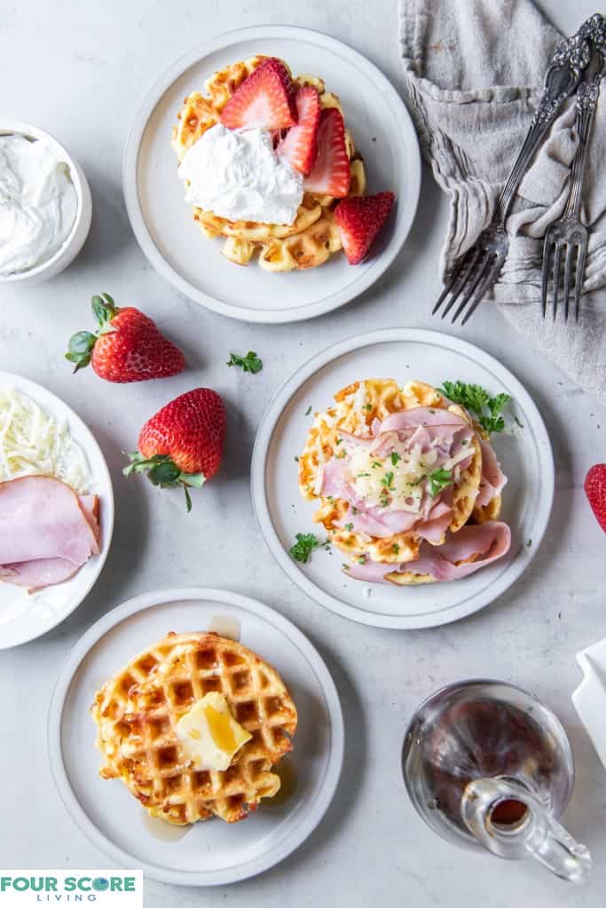 Aerial view of a three cooked keto waffles, each on a small plate, all in prominent full view one with fresh halved strawberries and whipped cream, one with sliced ham, melted white cheese and green garnish, one with a pat of butter and sugar free syrup, with a glass bottle of syrup in the right lower corner of the frame, with three whole strawberries, a natural kitchen towel, two forks, a small dish of whipped cream and a partial view of a small plate of ham and shredded cheese all on a light colored stone surface.