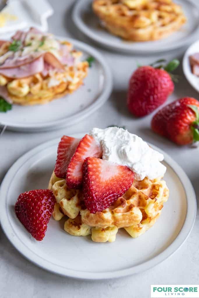 Aerial diagonal view of three cooked keto waffles, each on a small plate, one in full view with fresh halved strawberries and whipped cream, one in full view with and in soft focus with sliced ham, melted white cheese and green garnish, one in ¾ view with a pat of butter and sugar free syrup, with two whole strawberries all resting on a light stone surface.