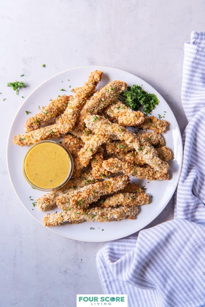 Aerial view of breaded and cooked chicken fries on a white plate with green herb garnish and a small dish of honey mustard dipping sauce with a light grey and white striped kitchen towel on a white stone surface.