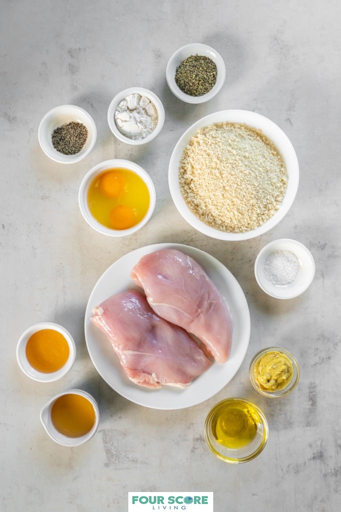 Aerial view of the ingredients in small and medium white dishes, to make chicken fries including bread crumbs, cracked raw eggs, two chicken breasts, seasonings, and flour and ingredients for honey mustard dipping sauce, honey, mustard, olive oil and apple cider vinegar.