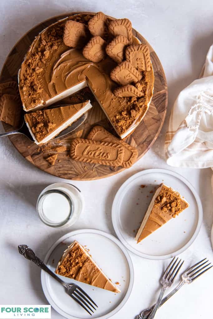 Aerial view of a sliced Biscoff cheesecake with two slices on small white plates, with three forks and a clear glass milk bottle and an ivory kitchen towel nearby.