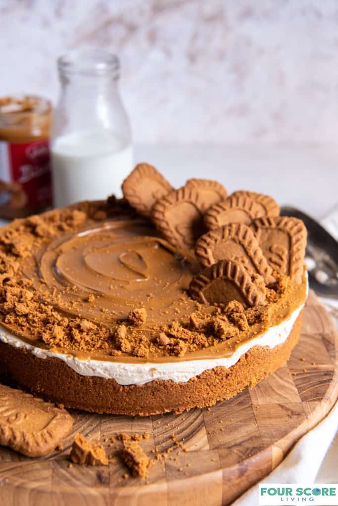Diagonal close up shot of Biscoff Cheesecake garnished with cookie crumbs and whole Biscoff cookies, placed on a round wooden cutting board with a jar of Biscoff spread and a glass bottle of milk in soft focus in the background, all resting on a white stone surface.