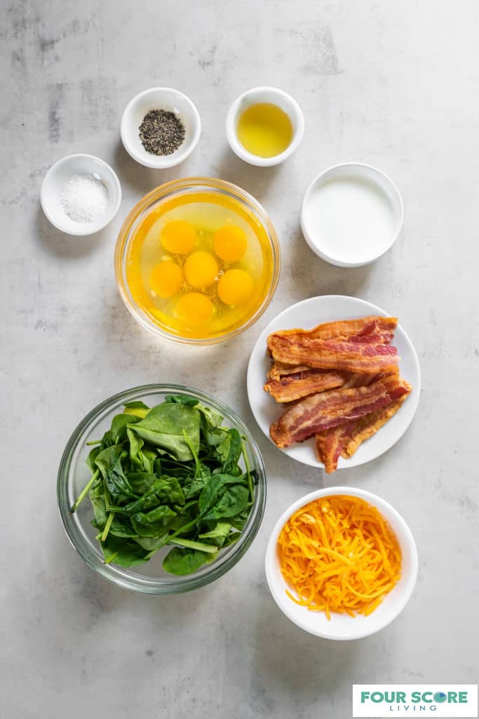 Aerial view of white and clear bowls each containing an ingredient for egg bites, including cooked bacon strips, shredded cheddar cheese, fresh spinach, cracked eggs, slat, pepper, oil and heavy cream all resting on a white stone surface.
