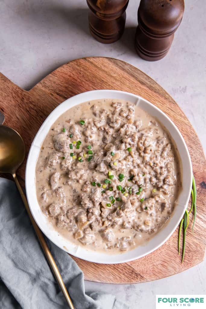 Aerial view of a white bowl of keto sausage gravy with chopped green onions sprinkled on top, layered on a warm colored wood cutting board with a large brass serving spoon, whole green onions and a light grey dishtowel, all resting on a white stone surface.
