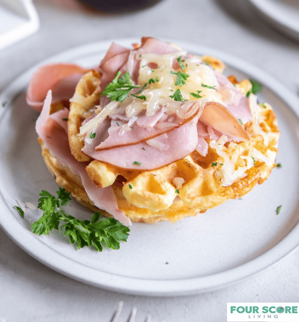 Diagonal closeup view of a keto chaffle layered with ham and melted white cheese and sprinkled with green garnish on a white plate resting on a white stone surface.