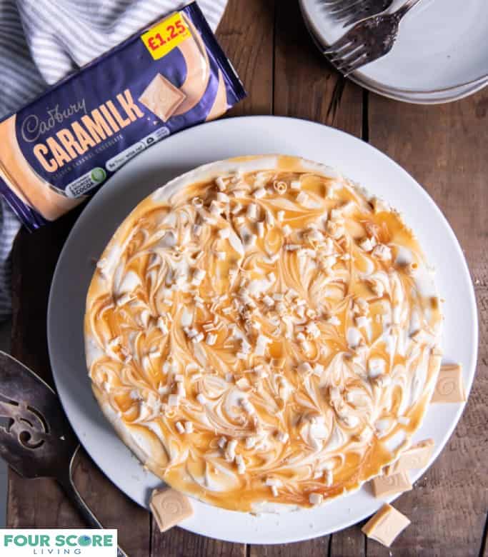 Aerial view of a caramilk cheesecake with caramel swirls and shaved caramilk chocolate garnish on a white plate with a packaged Cadbury caramilk bar, a pinstriped kitchen towel, two plates, two forks and a cake serving utensil all resting on a dark wooden surface.