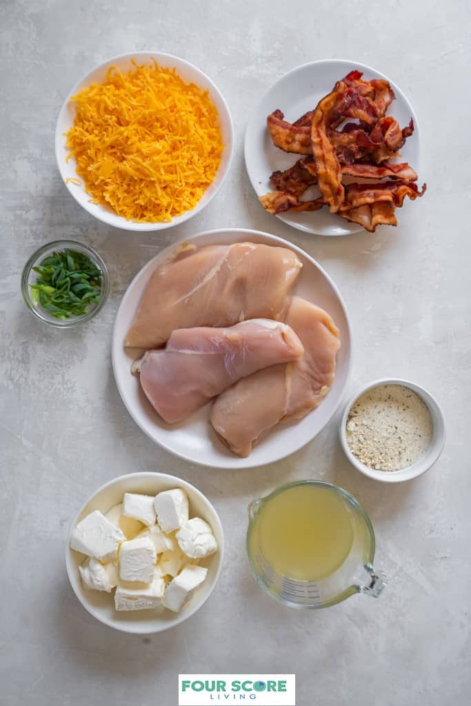 Aerial view of ingredients to make Instant Pot Crack Chicken on plates and bowls such as chicken breasts, cream cheese, ranch seasoning, cheddar cheese, bacon, onions, chicken broth all resting on a white stone surface.