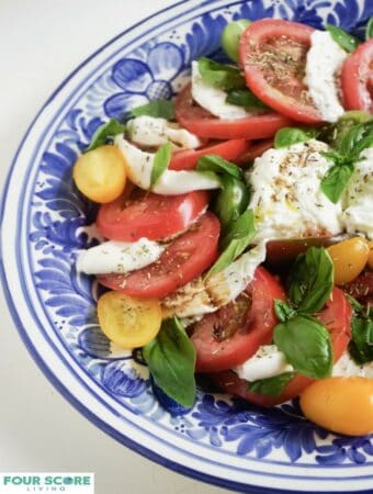 A burrata caprese with burrata cheese, sliced red tomatoes, sliced yellow Roma tomatoes, fresh basil, drizzled with oil and balsamic vinegar and sprinkled with fresh herbs on a blue plate.