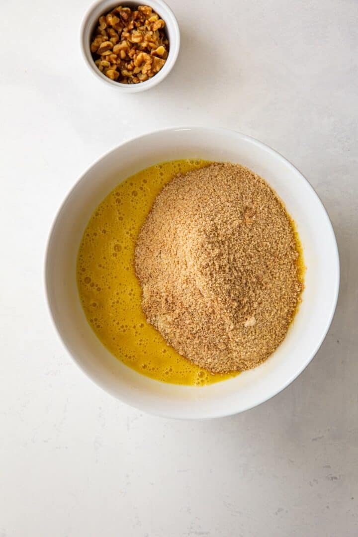 white bowl containing the wet and dry banana bread ingredients. Also a small white bowl with walnuts on a countertop.
