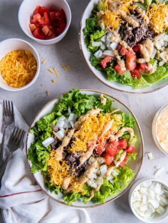 Two plates of salad greens containing cooked ground beef, shredded cheddar cheese, diced red tomatoes, diced white onions, drizzled big mac sauce and a sprinkle of sesame seeds.
