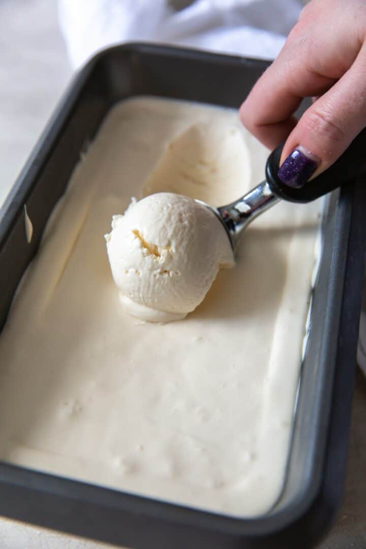 A feminine hand scooping ice cream with an ice cream scoop from a dark metal loaf pan.