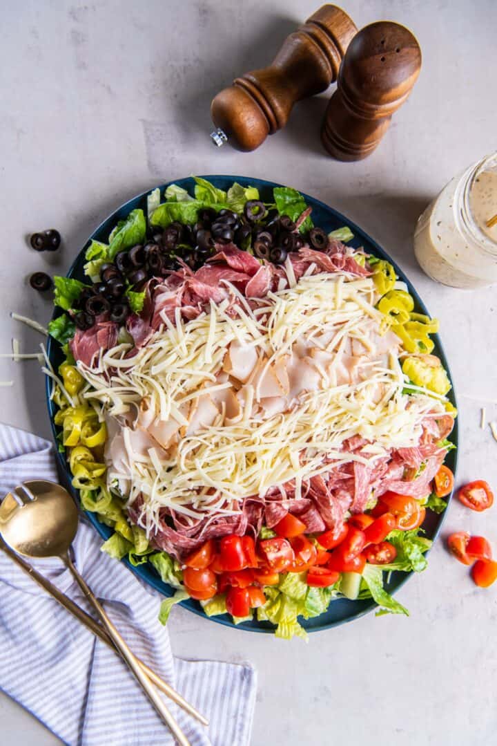 A fully prepared grinder salad on a plate, all ingredients layered including romaine lettuce, red cherry tomatoes, shredded cheese, sliced turkey, salami and capicola.