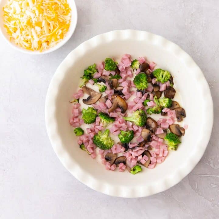 A bowl of shredded cheese and a pie pan with ham, mushrooms and broccoli.