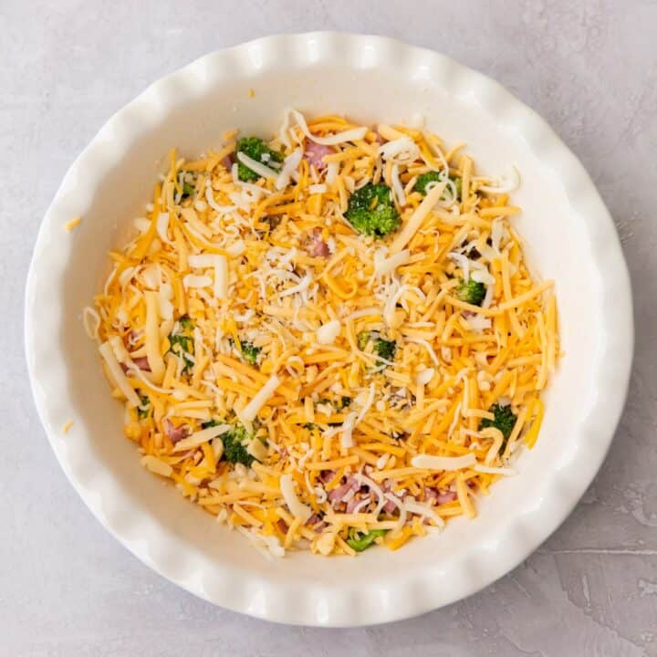A pie pan with shredded cheese layered on top of ham, broccoli and mushrooms.