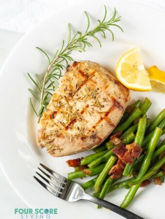 Grilled pork chop on a plate served with green beans, lemon wedges, and a rosemary garnish.