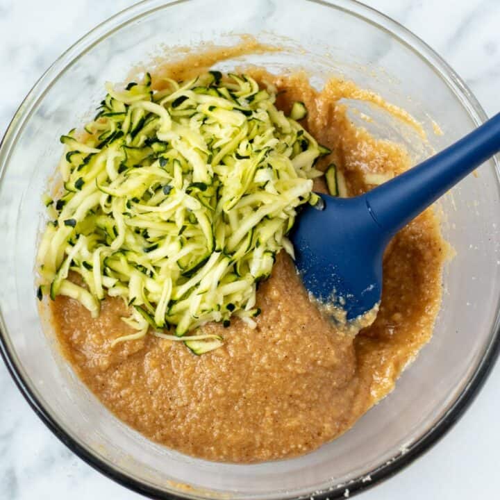 Shredded zucchini being added to the batter in a glass bowl.