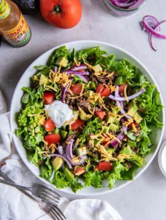 Taco salad in a bowl with chopped tomatoes, sliced red onion, diced avocado, cooked ground beef, shredded cheese and a dollop of sour cream.