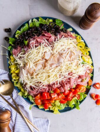 A fully prepared grinder salad on a plate, all ingredients layered including romaine lettuce, red cherry tomatoes, shredded cheese, sliced turkey, salami and capicola.