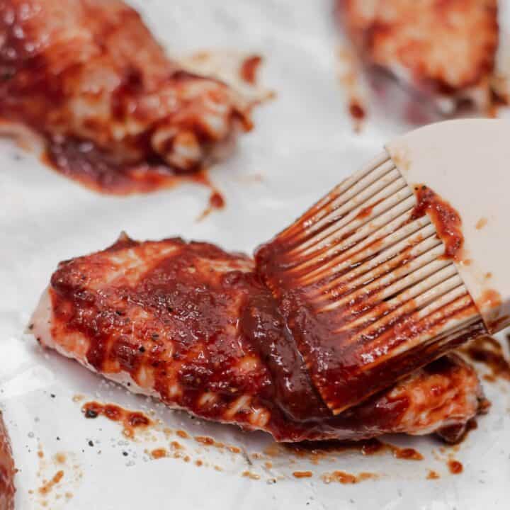 BBQ sauce being brushed onto raw chicken wings on foil lined baking sheet.