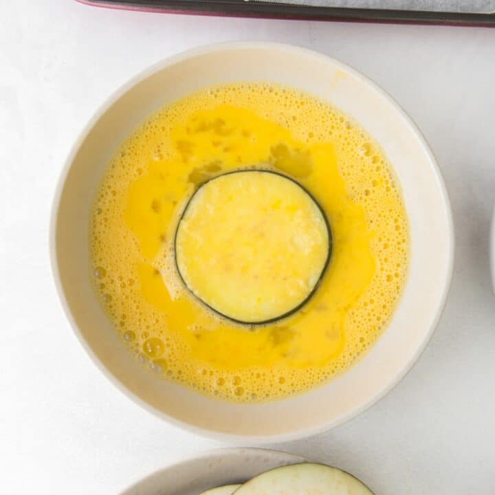 An almond flour coated eggplant round being dipped in egg.