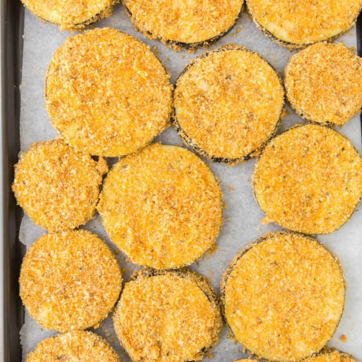 Breaded eggplant rounds on a parchment lined baking sheet, ready to go into the oven.