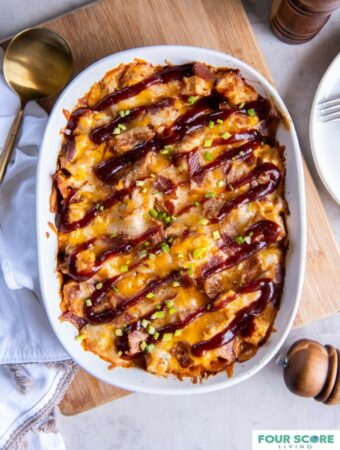 Oval casserole dish with a baked BBQ chicken casserole drizzled with BBQ sauce and set on a wooden cutting board next to a kitchen towel and a large round serving spoon.