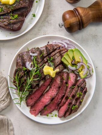 Two white plates of sirloin tip steak, one sliced and each garnished with sliced avocado, purple onion and a pat of butter.