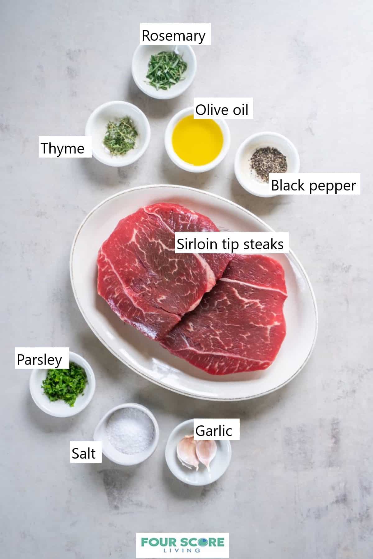 An oval platter with two raw sirloin tip steaks and small dishes of herbs, spices, salt, two garlic cloves and olive oil.