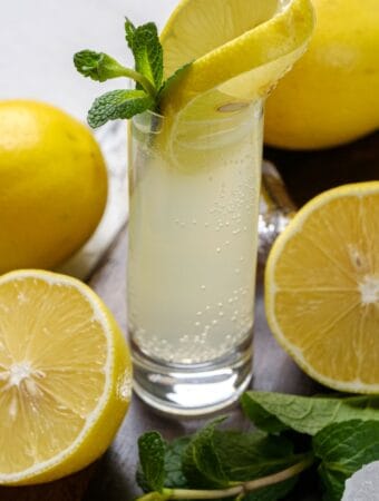 White Tea Shot in a wooden cutting board. There is a slice of lemon and sprig of mint in the shot, and the glass is surrounded by mint, lemons, and ice.