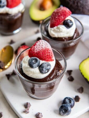 2 glass dessert cups filled with chocolate avocado pudding. The pudding is topped with a dollop of whipped cream, a half of a strawberry and two blueberries.